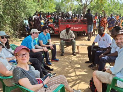 Reisegruppe sitzt mit afrikanischen Männern im Kreis, dahinter eine Gruppe von Menschen hinter einem kleinen Truck.